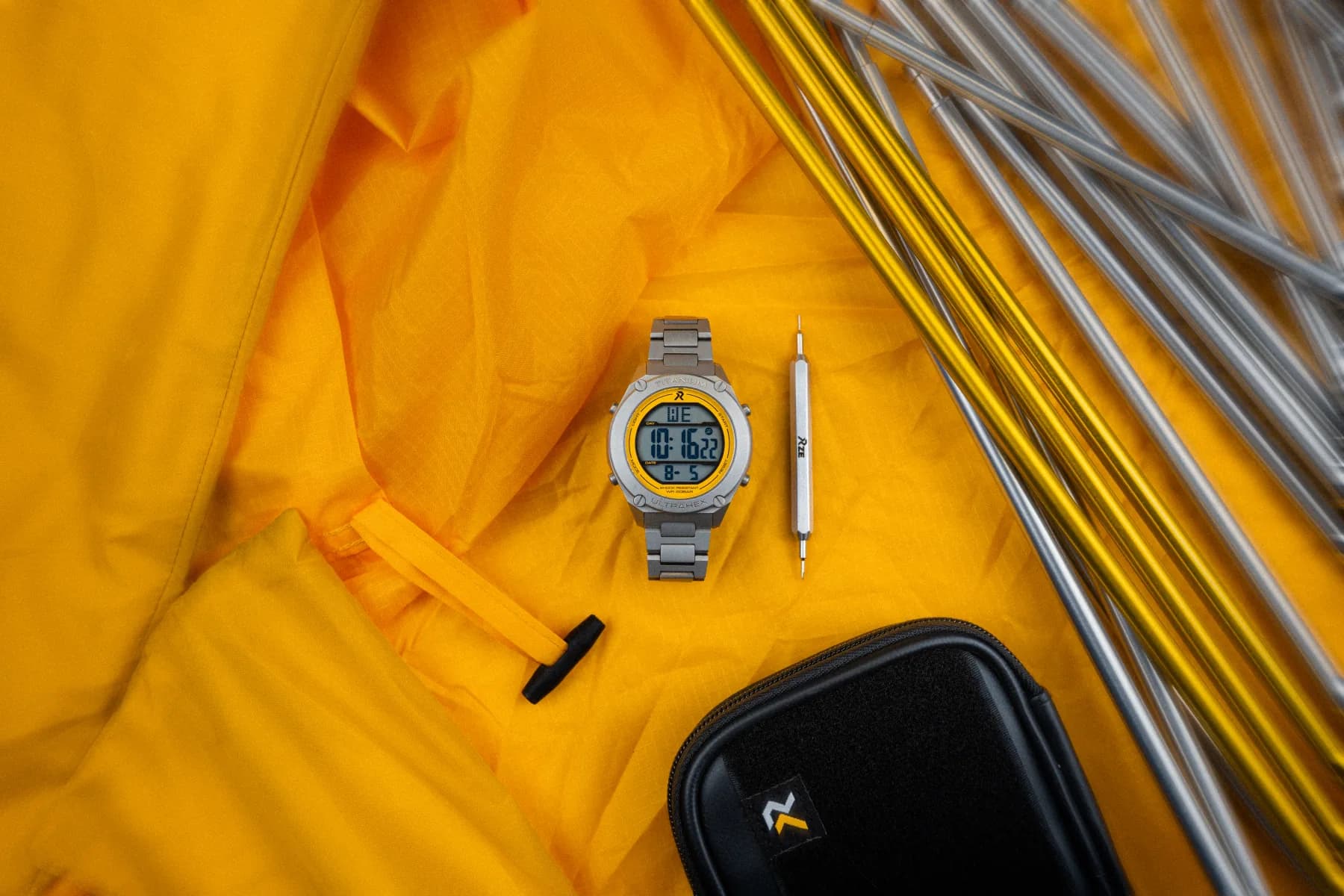 RZE titanium watch laid on a yellow tent fly beside tent poles, spring bar tool and travel case