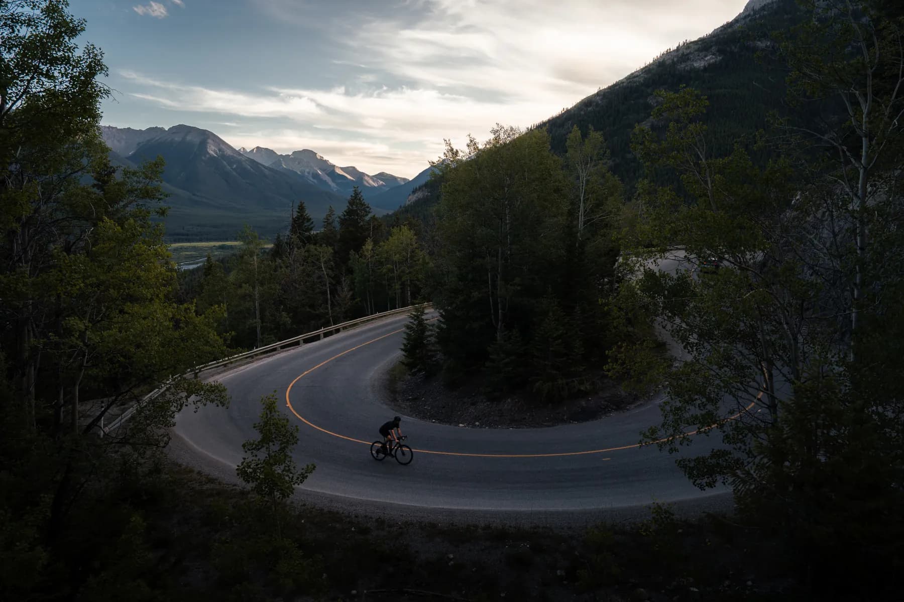 Lone cyclist carving a hairpin bend on a Canadian Rockies mountain road at dusk